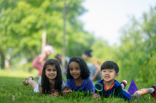 A small group of three elementary children lay in the grass with magnifying glasses as they pose for a portrait.  They are each dressed casually and smiling as they enjoy taking their science lesson outside.