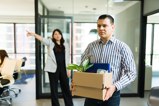 Depressed mid adult male employee holding box of belongings being dismissed by angry boss in office