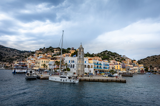 Symi, Greece - May 30, 2023:  Colorful architecture  and a famous clock tower on the sea coast of Symi island, Greece. Sailing boat moored at the pier.