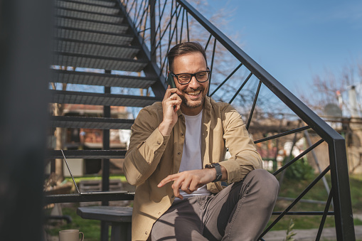 One man caucasian male sit on the steps outdoor while taking a brake from work with laptop use mobile phone to make a call talking happy smile confident real person