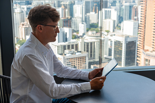 Portrait of young European businessman in glasses using tablet computer in panoramic office. Concept of communication and social media