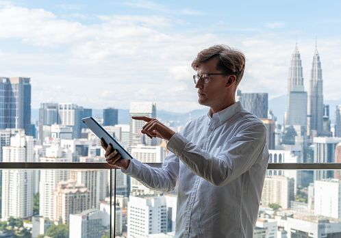 Portrait of serious young European businessman in white shirt using tablet computer standing near panoramic window. Concept of communication
