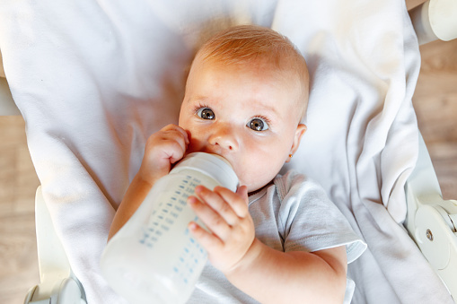 Cute little newborn girl drinking milk from bottle and looking at camera on white background. Infant baby sucking eating milk nutrition lying down on crib bed at home. Motherhood happy child concept