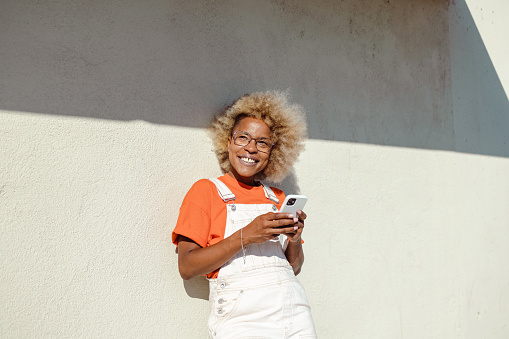 African American woman with afro dyed hair posing in front of the wall and using smart phone