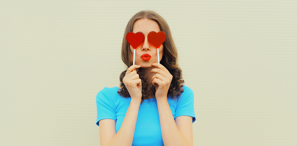 Portrait of beautiful young woman covering her eyes with red heart shaped lollipop blowing her lips with lipstick sending sweet air kiss