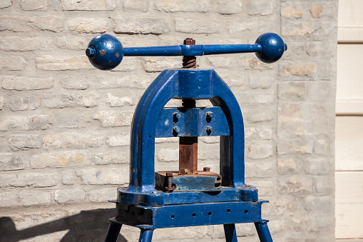 Picture of a manual printing press on display on a brick wall. A printing press is a mechanical device for applying pressure to an inked surface resting upon a print medium, thereby transferring the ink.