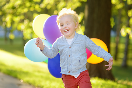 Little boy having fun on celebrating birthday party. Happy child with bunch of bright multicolor balloons. Preschoolers or toddlers baby birthday party in sunny park. Summer outdoor festival for kids