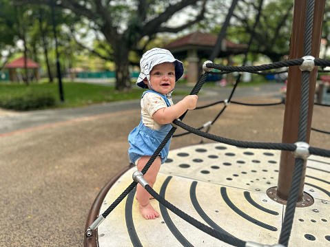 Multiracial 0-11 month old baby boy smiles as he holds onto the rope at the playground, helping himself stand up.