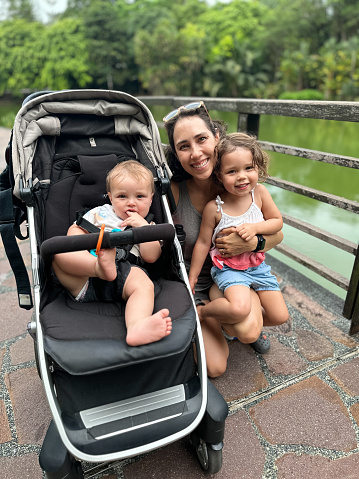 Eurasian mother and her multiracial baby son and toddler daughter smile as they pose for a picture while walking on a trail through a garden in Singapore.