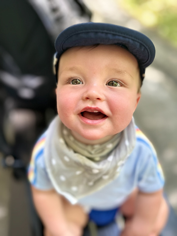 Multiracial, 0-11 month old baby boy smiles up at camera while sitting on a bench in a public park.