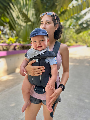 Multiracial, 0-11 month old baby boy giggles and smiles as his Eurasian mother carries him through a public botanical garden while traveling in Singapore.