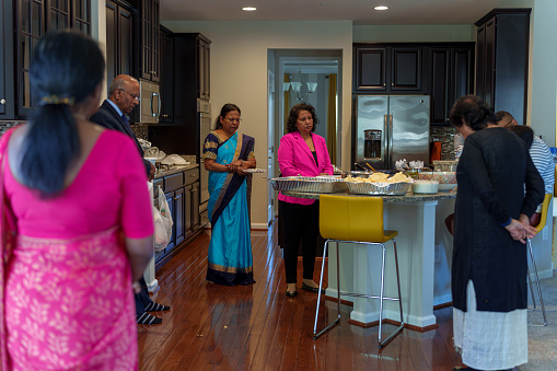 Multi-generation Indian family gathers in the kitchen at home and prays before eating together at a casual family celebration.