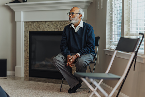 Senior Indian male sits on a folding chair in the living room, chatting during a family gathering.