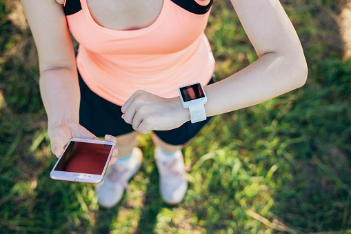 From a shoulder-level perspective,An Asian woman finishes her outdoor fitness training and carefully compares the data on her phone and watch, including distance, speed, heart rate, and calorie expenditure, to ensure the effectiveness of her workout and achieve her fitness goals.