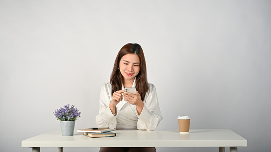 A happy and charming young Asian businesswoman or female office worker using her smartphone at her desk. isolated white background, text, chat, message, social media, mobile application