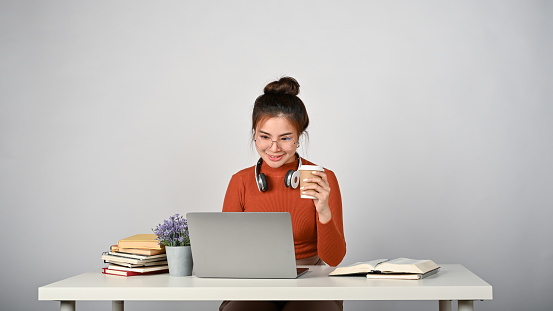 Young adorable Asian student working with laptop computer while holding a coffee cup in her hand, white isolated background.