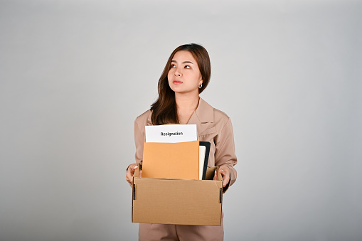 Sad young businesswoman holding a box full of her stuff, feeling sad after quitting her job, white isolated background.