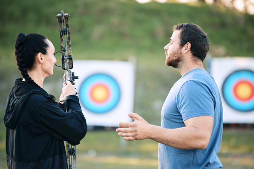 Sports, archery coach or bow and arrow learning for archer competition, athlete challenge or girl training practice. Teacher, teaching talk or man coaching woman on precision, aim and target shooting
