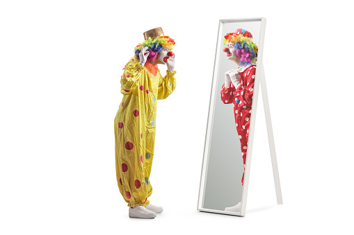 Clown in a yellow costume standing in front of a mirror and looking at a clown in a red costume isolated on white background