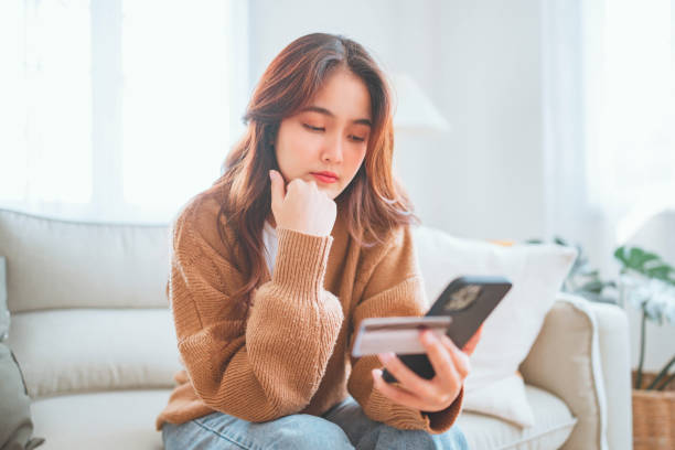 Young asian woman problems with paying, Sad female internet shopper sit on couch hold phone and credit card with feeling depressed and worry for distress suffer of overspending money from card account mujer asiática joven problemas con el pago, triste compradora de internet sentarse en el sofá sostener el teléfono y la tarjeta de crédito con sentirse deprimida y preocuparse por la angustia sufrir de gastar demasiado dinero de la cuenta de la tarjeta