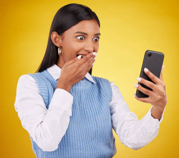 wow, surprise and woman with phone in studio space reading text or email on yellow background. technology, communication and good news, happy indian girl with social media gossip or online content. - young adult reading newspaper the media imagens e fotografias de stock