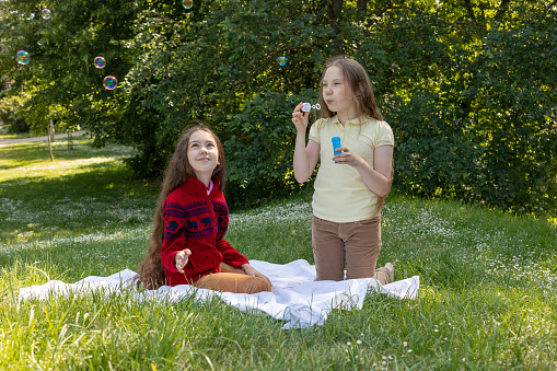 Cheerful Little Asian Caucasian Girls Of Elementary School Age Blowing Soap Bubbles In Summer Park, Meadow. Happy Childhood. School Break Time. Earth Day. Children Protection Day. Horizontal Plane.