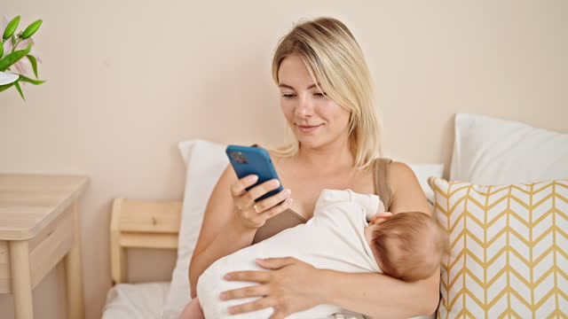 Mother and daughter sitting on bed breastfeeding baby using smartphone at bedroom