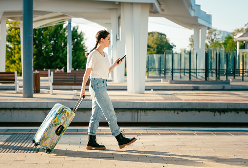 Side view of young woman walking with her luggage and using cellphone. Concept of modern travel and vacation.