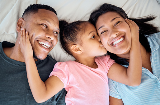 Happy family, kid kiss and bed with a girl, mother and father with a smile, love and parent care. Happiness, hug and black family in a bedroom lying relax together in a morning with a child at home