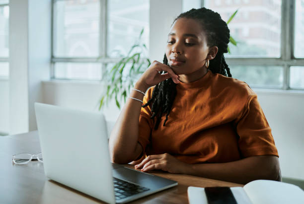 une jeune femme d’affaires noire travaillant sur son ordinateur portable - main au menton photos et images de collection