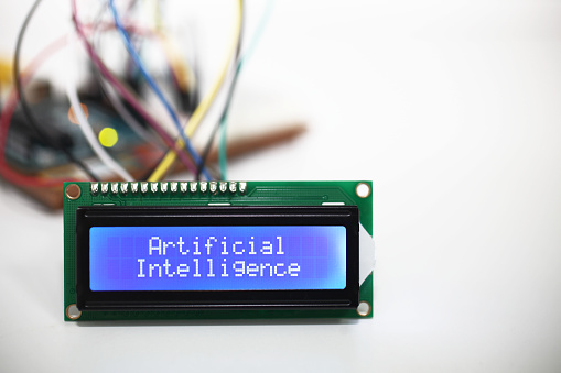 A blue LCD display showing the words 'Artificial Intelligence' with wires coming from a computer circuit board in the background.