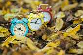 Daylight saving time, fall back concept with blue, yellow, red alarm clocks and autumn leaves foliage, selective focus.