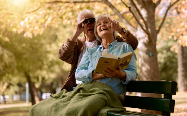 Park, bench and binoculars with a senior couple birdwatching together outdoor in nature during summer. Spring, love and book with a mature man and woman bonding while sitting in a garden for the view parque, banco y binoculares con una pareja mayor observando aves juntos al aire libre en la naturaleza durante el verano. primavera, amor y libro con un hombre y una mujer maduros que se unen mientras están sentados en un jardín para la vista
