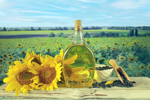 A bottle of sunflower oil, fresh sunflower flowers and seeds on the table, on the background of a landscape with a blooming field, summer, autumn