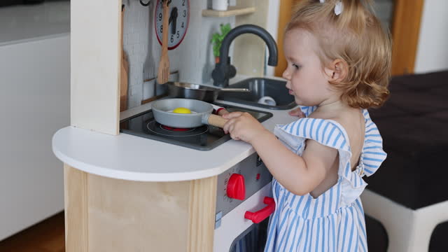 Cute little baby girl playing on toy kitchen at home, pretends frying eggs