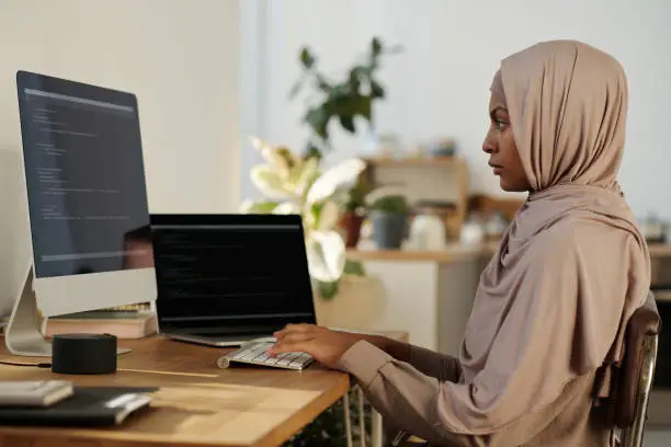 Young Muslim female webdesigner or programmer in beige headscarf typing on computer keyboard and looking at screen during network