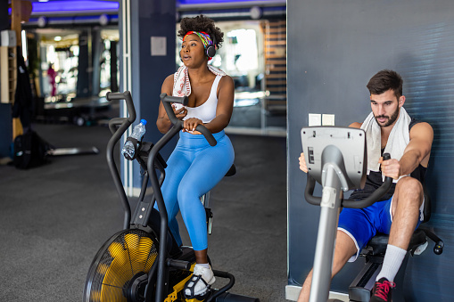 A female of African-American ethnicity is doing exercise training in a gym using a bike while her male friend is resting.