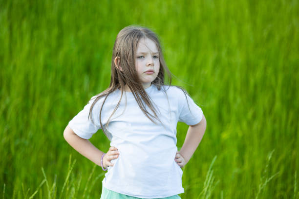 A young girl with long blonde hair lies on a green lawn, her head supported by her hand as she thoughtfully examines a blade of grass. She wears a brown quilted jacket, and her serene expression captures a moment of quiet reflection. The backdrop of trees and blurred background elements add to the tranquil and contemplative atmosphere of this outdoor scene.
