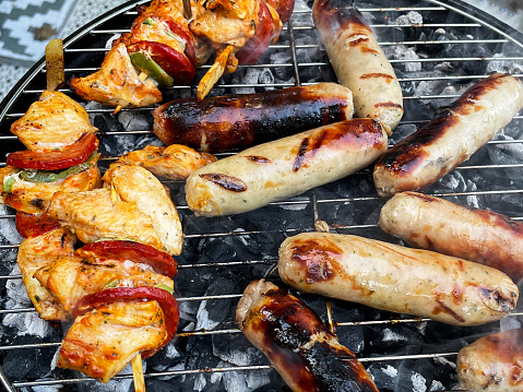 Stock photo showing close-up, elevated view of a portable kettle charcoal barbecue in a garden setting, stood on a wooden patio table featuring a large expanse of white, interconnecting, white plastic decking tiles in the background. Pictured cooking on the barbecue are some sausages and kebabs on wooden skewers made from chicken pieces and chorizo slices.