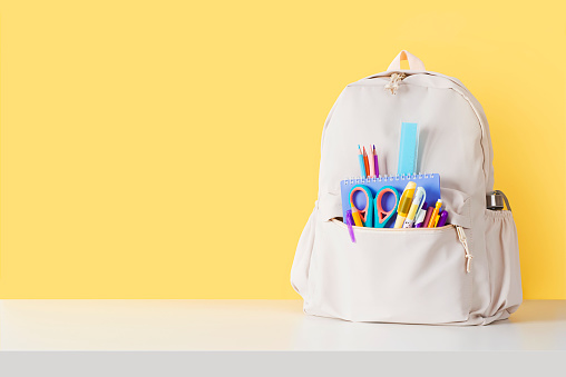 Backpack with school supplies on white table near yellow wall