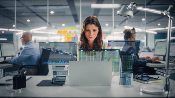 joven empresaria feliz usando computadora portátil en una oficina moderna con colegas. elegante y hermoso asesor financiero trabajando. holograma vfx edit visualización de la interfaz de la bolsa de valores, gráficos abiertos. - stock market analyzing computer finance fotografías e imágenes de stock