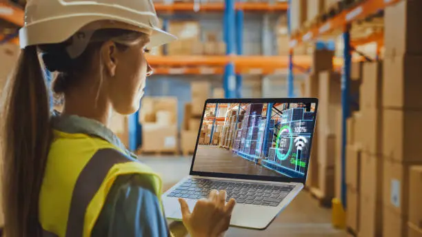 Futuristic Technology Warehouse: Female Worker Doing Inventory, Using Augmented Reality Program On Laptop Computer. Woman Analyzes Digitalized Products Delivery Infographics in Distribution Center.