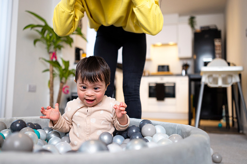 A young male toddler who has down syndrome is sitting in a ball pit on the living room floor, he is playing with the plastic balls at his home in Ashington, England. He is wearing a babygro and his mother is behind him.