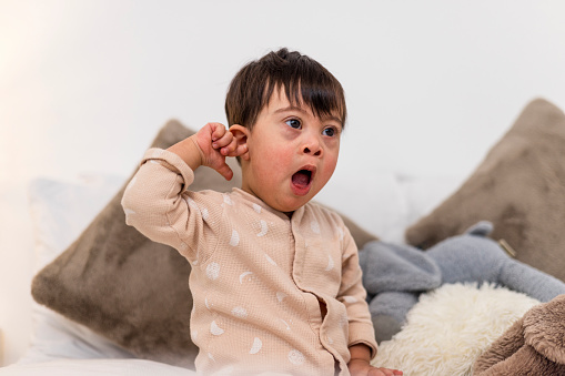 Front-view of a young male toddler who has down syndrome sitting on a bed at his home in Ashington, England. He is wearing a babygro and has cuddly toys by his side. He has his finger in his ear and he is yawning.