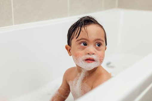 Front-view of a young male toddler who has down syndrome having a bubble bath in the bathtub at his home in Ashington, England. He is looking away from the camera.