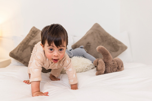 Front-view of a young male toddler who has down syndrome crawling on a bed at his home in Ashington, England. He is wearing a babygro and has cuddly toys by his side.