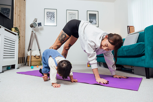 Young mother performing a yoga pose on her yoga mat in her living room at her home in Ashington, England. Her young male toddler who has down syndrome is with her having fun and copying her yoga pose, he is wearing dungarees.