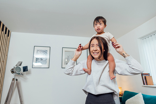Waist up of a young mother standing in her living room, her young male toddler son who has down syndrome is sitting on her shoulders, he is smiling and having fun with his mother, they are at their home in Ashington, England.