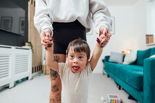 Close-up of a young male toddler who has down syndrome standing on the living room floor playing with his mother, they are holding hands at his home in Ashington, England. He is wearing a babygro and learning to walk, he is looking past the camera.