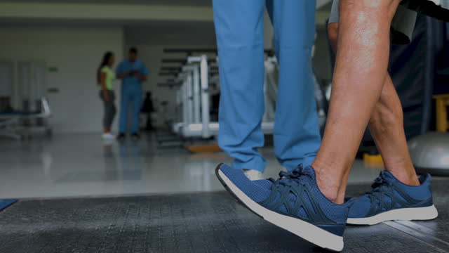 Close-up of unrecognizable man in physical therapy taking his first steps while therapist is standing next to him